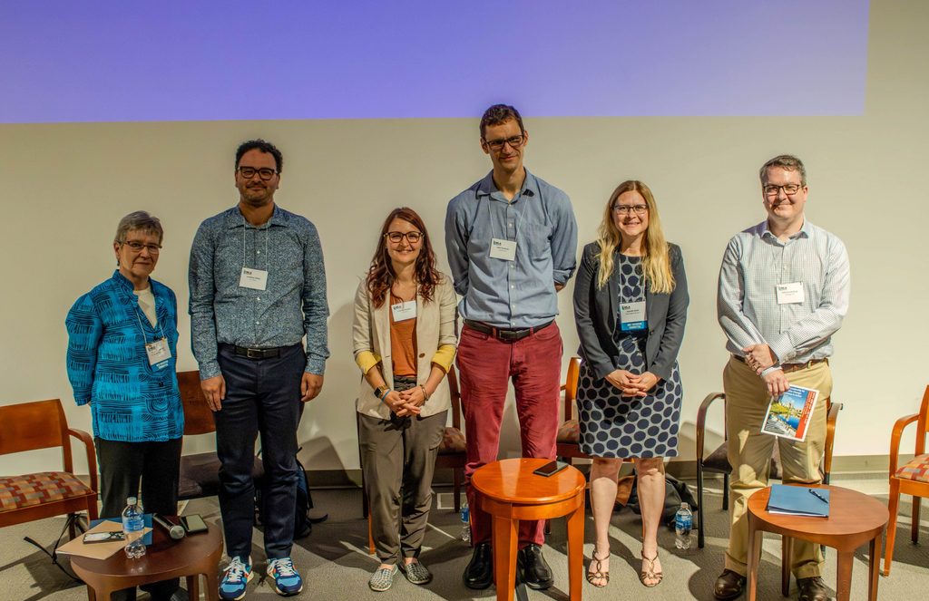 Six people pose in a row in front of a projector screen.
