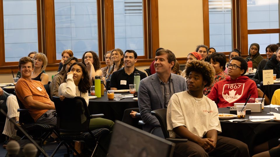 A crowd of people sitting at tables in Weitz 236 turn to look at something off-camera.