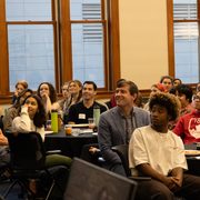A crowd of people sitting at tables in Weitz 236 turn to look at something off-camera.