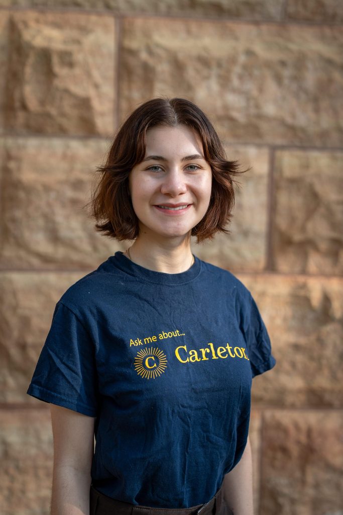 Portrait photo of Arielle Szycher standing against a wall in a Carleton shirt