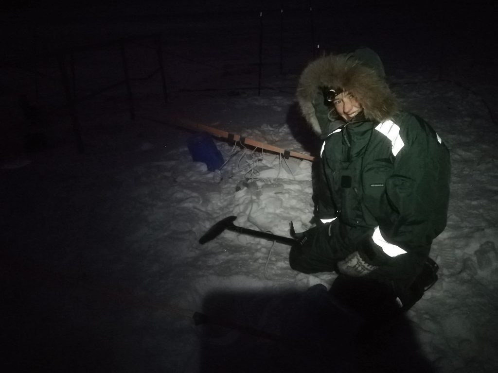 Torstenson wearing a headlamp and winter coat looks at the camera while kneeling in the snow with a pickaxe.