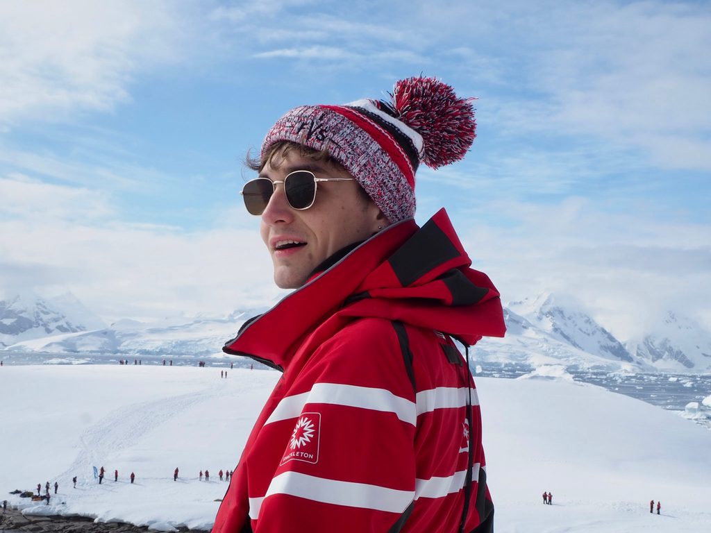 A portrait of Mitch Porter in sunglasses, a winter hat and coat standing in front of an ice field.