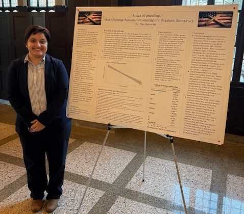 A student stands and smiles next to their comps poster in the Great Hall.