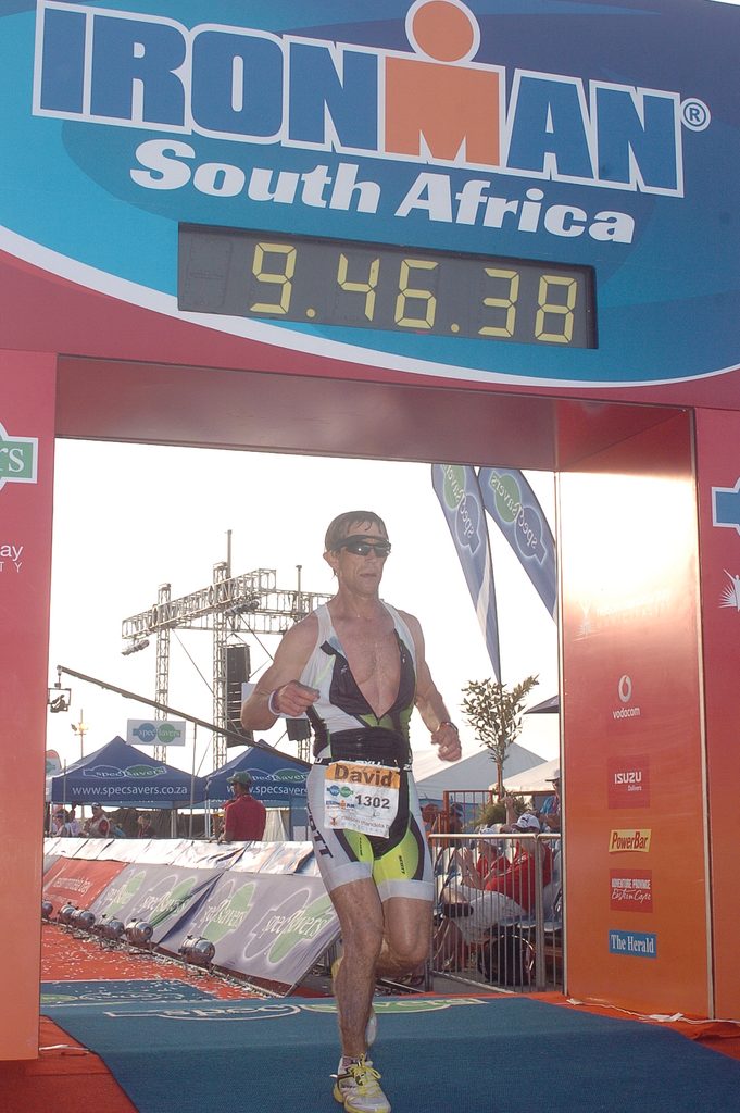 David Evans ’82 runs across the finish line under a sign reading, "Ironman South Africa."