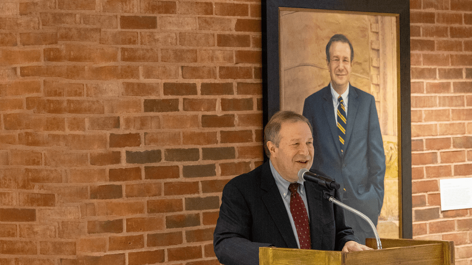 Steve Poskanzer giving a speech in front of his official presidential portrait.