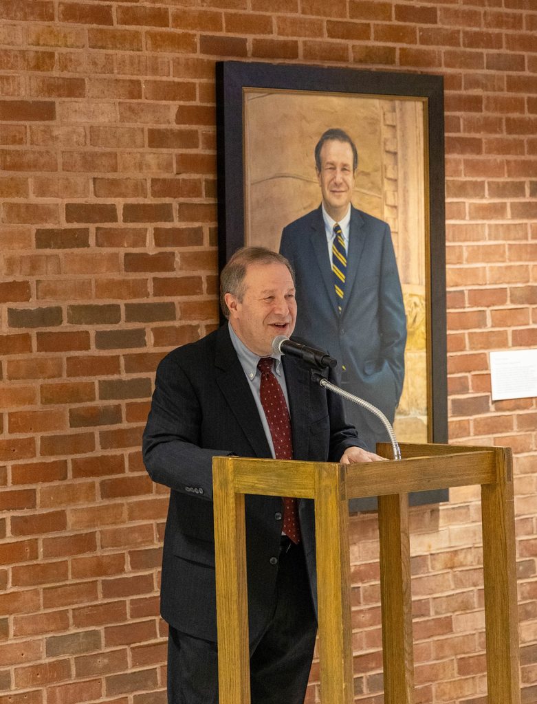 Steve Poskanzer giving a speech in front of his official presidential portrait.