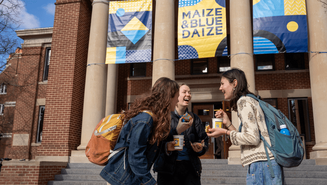 Three students laugh together in front of Sayles, which is decorated with Maize & Blue Daize banners.