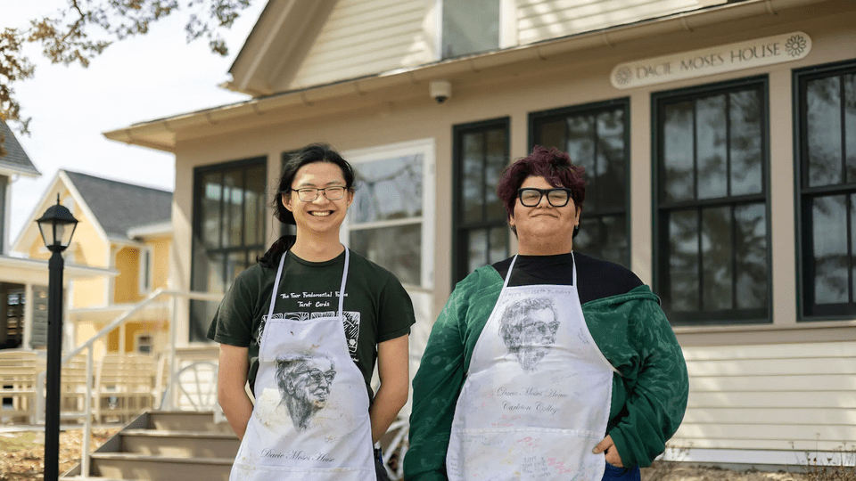 Two students in Dacie Moses aprons pose and smile in front of the house.