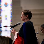 Provost Michelle Mattson clapping at the lectern on the Chapel stage, seen from the side with stained glass windows in the background.