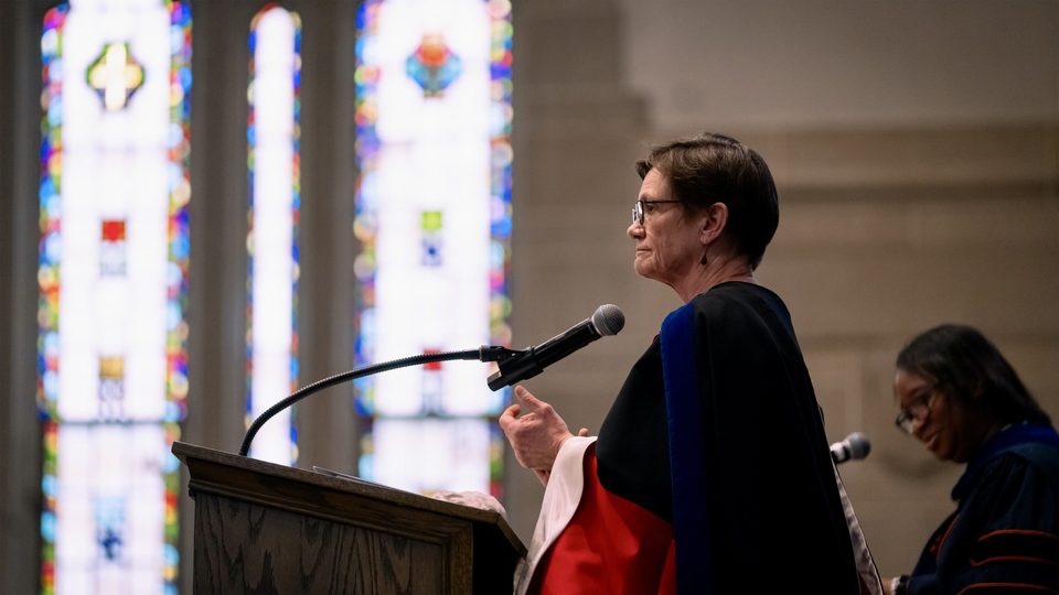 Provost Michelle Mattson clapping at the lectern on the Chapel stage, seen from the side with stained glass windows in the background.