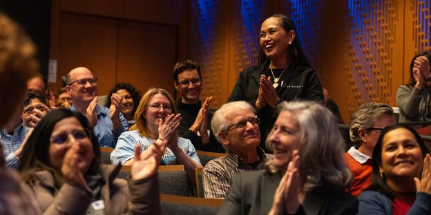 Phoebe Wilmot stands and smiles in Kracum Hall as surrounding seated staff and faculty applaud her.