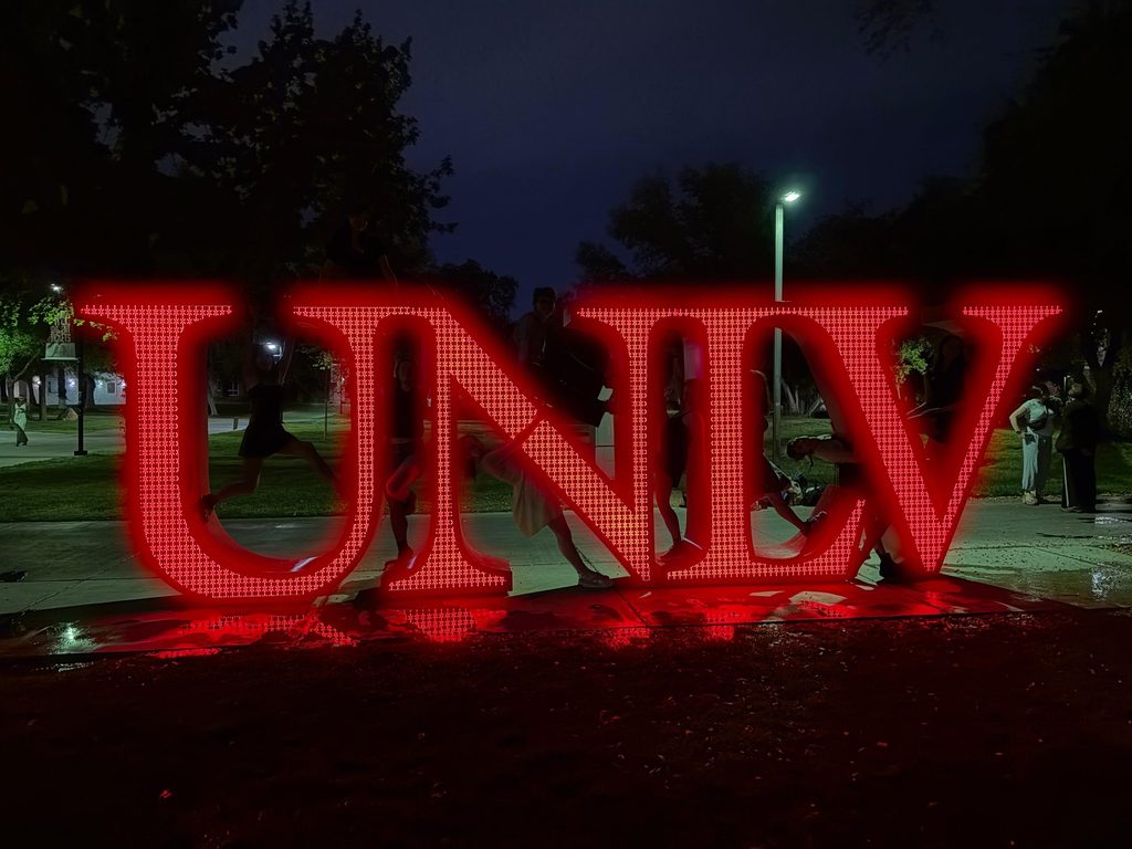 People lean on a bright red light-up LED sculpture of the letters UNLV at night.