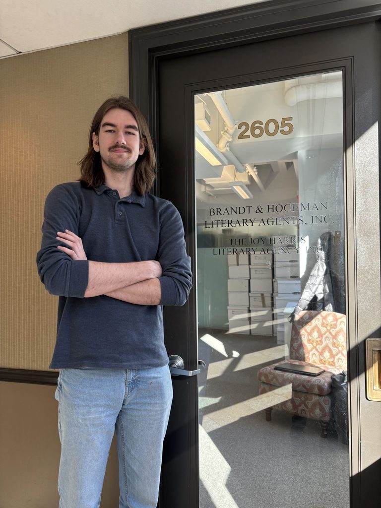 A student poses with arms crossed in front of the front door of a literary agency office.