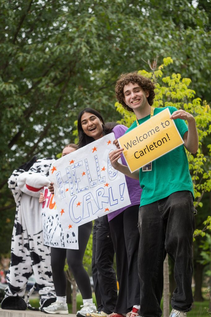 Students smile on the Carleton welcome sign with signs for New Student Week.