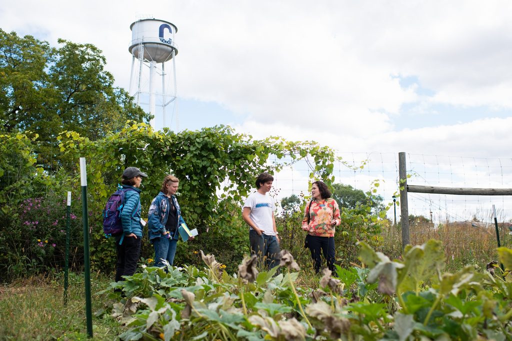 A woman and three students talk to each other near a community garden with a water tower in the background.