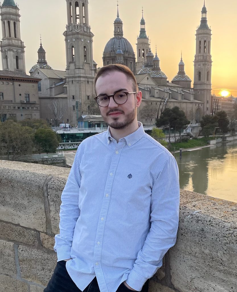 Marco on a bridge with a river and a building and sunset in the background.