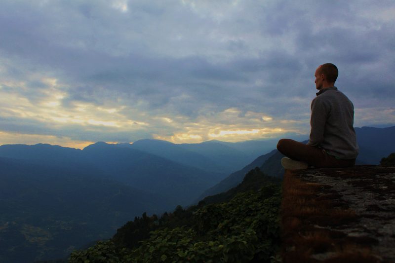 A man sits on the edge of a mountain, looking at the sunset