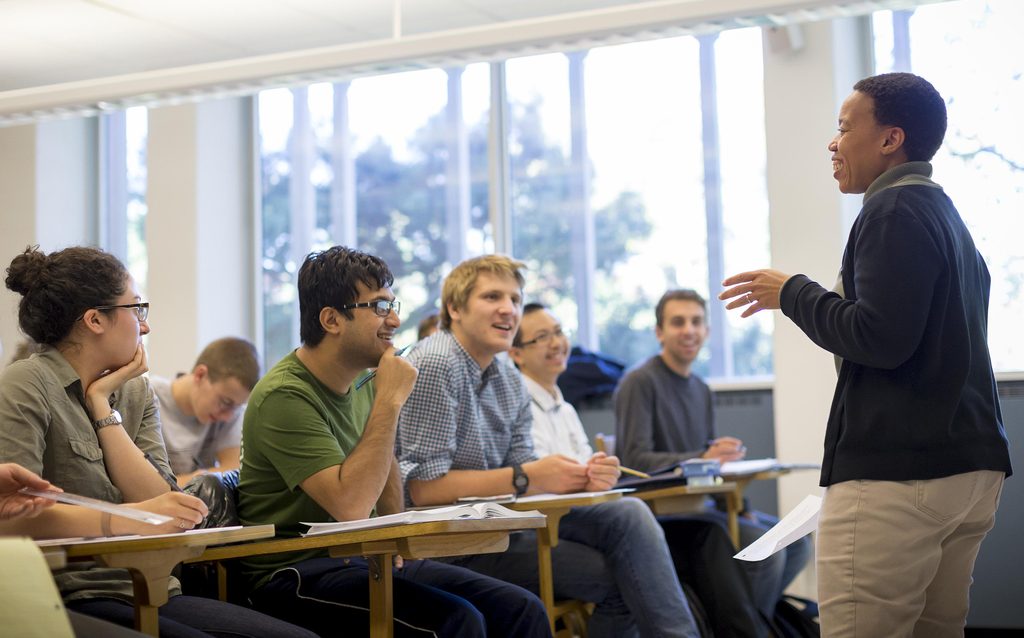 A professor stands and reads aloud from a paper in a classroom