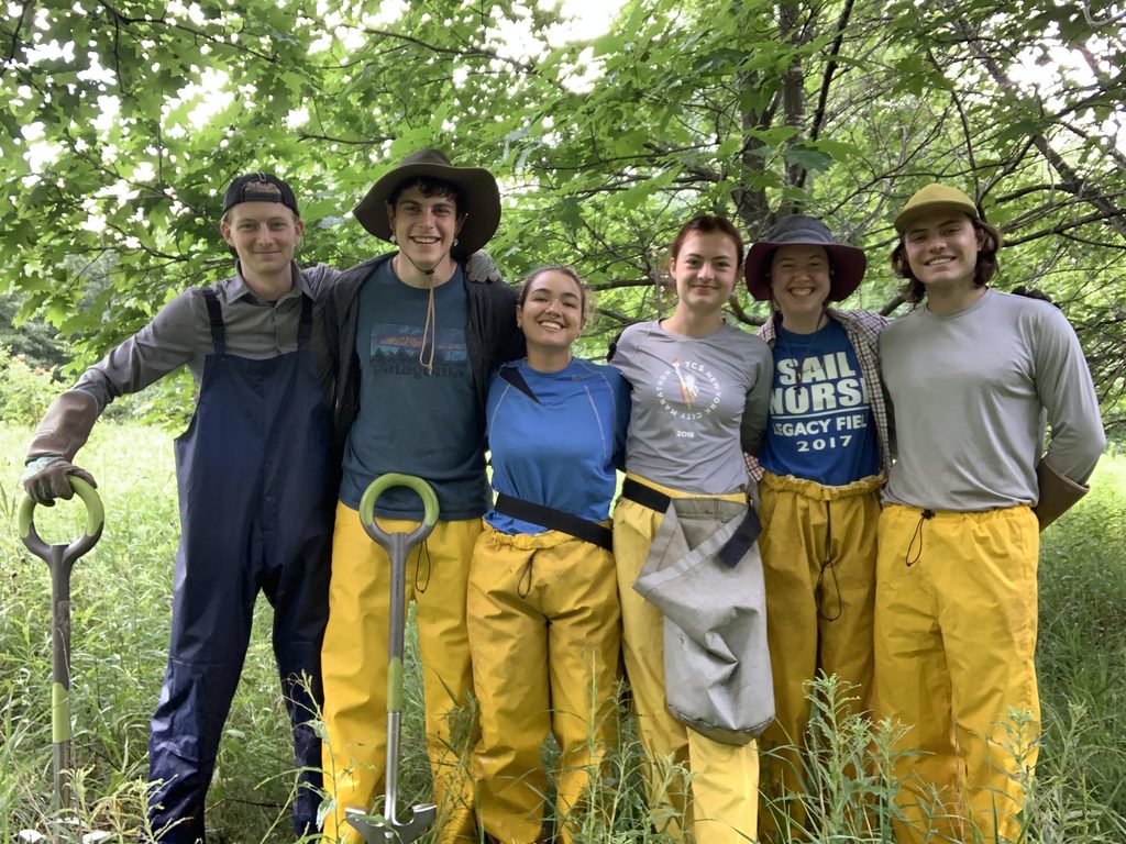 Group of six students in rain gear and hats in the forest to demonstrate student crew cohesion and enjoyment of working together.
