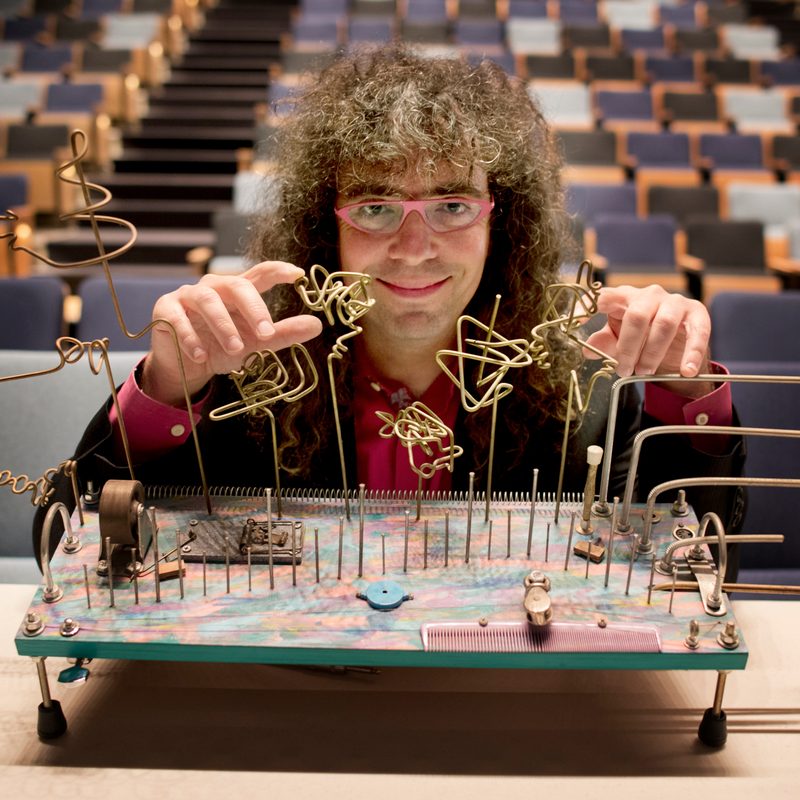 A man with long curly hair and red glasses smiles and poses in front of a multicolored handmade instrument made from found junk and hardware like twisted wires, nails and a moustrap