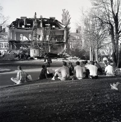 students sit on a hillside watching a building being torn down