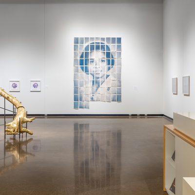 Installation image of the faculty show. Jade Hoyer's large installation of prints that come together to create an image of her mother as a nurse sits in the center of the image. Stephen's sculpture, a gold covered segmented tree on stilts, is to the left and in front of Jade's work. To the right there are four prints by Eleanor Jensen framed in wood. These prints are too far away to see the details. In the foreground and bottom right corner of the image you see the front of the museum reception desk.