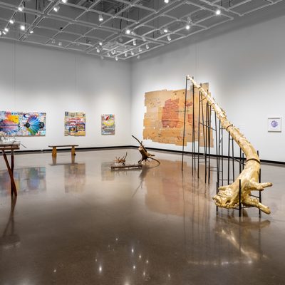 Large white wall exhibition space with three tree trunk sculptures in the center covered in gold and silver: one is extending into the ceiling segmented and supported on silts, the second smaller piece is resting on a hand made table with irregular branch legs, and the third installation is a selection of smaller branches gathered on the floor, all created by Stephen Mohring. The walls lining the room, from left to right, are four collaged panel paintings by David Lefkowitz, a collage of cardboard boxes flattened onto the wall into an irregular grid with a painting of a cargo shipping boat by David Lefkowitz, and three white framed prints by jade Hoyer.