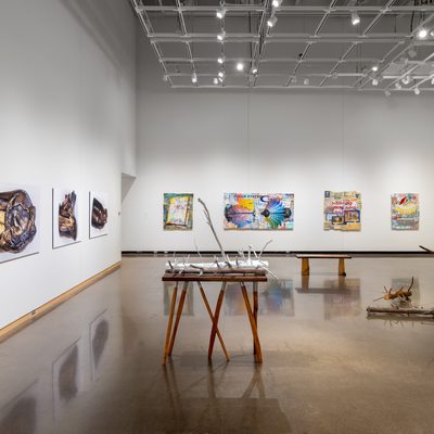 Large white wall exhibition space with two tree trunk sculptures in the center covered in gold and silver: one is extending into the ceiling segmented and supported on silts, the second smaller piece is resting on a hand made table with irregular branch legs, and the third installation is a selection of smaller branches gathered on the floor, all created by Stephen Mohring. Inthe distance, also in the center there is a handmade bench siting in front of the paintings on the far wall also created by Stephen. To the left of the sculptures are four large scale photographs of crushed and shattered bullets mounted on foam core created by Xavier Tavera. To the right of the photographs are four colorful collage panel paintings by David Lefkowitz with various realistic floating images painted on the surface. In this photo, to the far right, you catch a glimpse of David's cardboard box collage painting on the wall containing a painting of a red cargo boat.