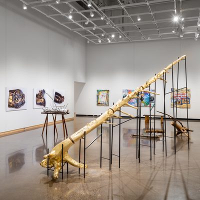Large white wall exhibition space with three tree trunk sculptures in the center covered in gold and silver: one is extending into the ceiling segmented and supported on silts, the second smaller piece is resting on a hand made table with irregular branch legs, and the third installation is a selection of smaller branches gathered on the floor, all created by Stephen Mohring. To the left and behind the sculptures are four large scale photographs of crushed and shattered bullets mounted on foam core created by Xavier Tavera. To the right of the photographs are three colorful collage panel paintings by David Lefkowitz with various realistic floating images painted on the surface.
