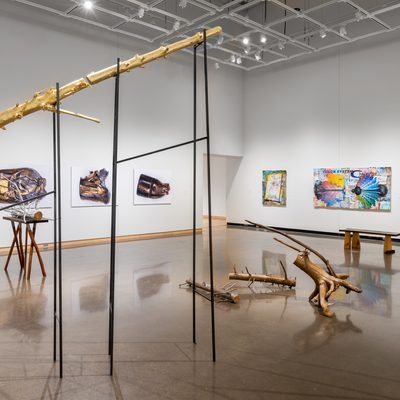 Large white wall exhibition space with three tree trunk sculptures in the center covered in gold and silver: one is extending into the ceiling segmented and supported on silts, the second smaller piece is resting on a hand made table with irregular branch legs, and the third installation is a selection of smaller branches gathered on the floor, all created by Stephen Mohring. To the left and behind the sculptures are four large scale photographs of crushed and shattered bullets mounted on foam core created by Xavier Tavera. To the right of the photographs are three colorful collage panel paintings by David Lefkowitz with various realistic floating images painted on the surface.