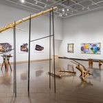Large white wall exhibition space with three tree trunk sculptures in the center covered in gold and silver: one is extending into the ceiling segmented and supported on silts, the second smaller piece is resting on a hand made table with irregular branch legs, and the third installation is a selection of smaller branches gathered on the floor, all created by Stephen Mohring. To the left and behind the sculptures are four large scale photographs of crushed and shattered bullets mounted on foam core created by Xavier Tavera. To the right of the photographs are three colorful collage panel paintings by David Lefkowitz with various realistic floating images painted on the surface.