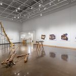 Large white wall exhibition space with three tree trunk sculptures in the center covered in gold and silver: one is extending into the ceiling segmented and supported on silts, the second smaller piece is resting on a hand made table with irregular branch legs, and the third installation is a selection of smaller branches gathered on the floor, all created by Stephen Mohring. To the left and behind the sculptures are four large scale photographs of crushed and shattered bullets mounted on foam core created by Xavier Tavera.