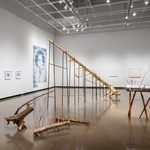 In the foreground of this photo Stephen's wooden sculptures are spread throughout the floor spacecovered in gold and silver: one is extending into the ceiling segmented and supported on silts, the second smaller piece is resting on a hand made table with irregular branch legs, and the third installation is a selection of smaller branches gathered on the floor. Behind these, from left to right, are three framed prints and a large scale print installation of a figure's headshot all created by Jade Hoyer, and to the right there are four framed prints on white paper by Eleanor Jensen.