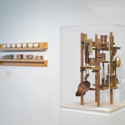 Two small shelves of ceramic cups by Kelly Connole. The top shelf contains white cups and the bottom contains irregularly earth colored woodfired cups. To the right of these shelves, on top of a pedestal, there is a clear vitrine of various hand forged spoons and cups suspended on a wooden display all created by Danny Satthoff.