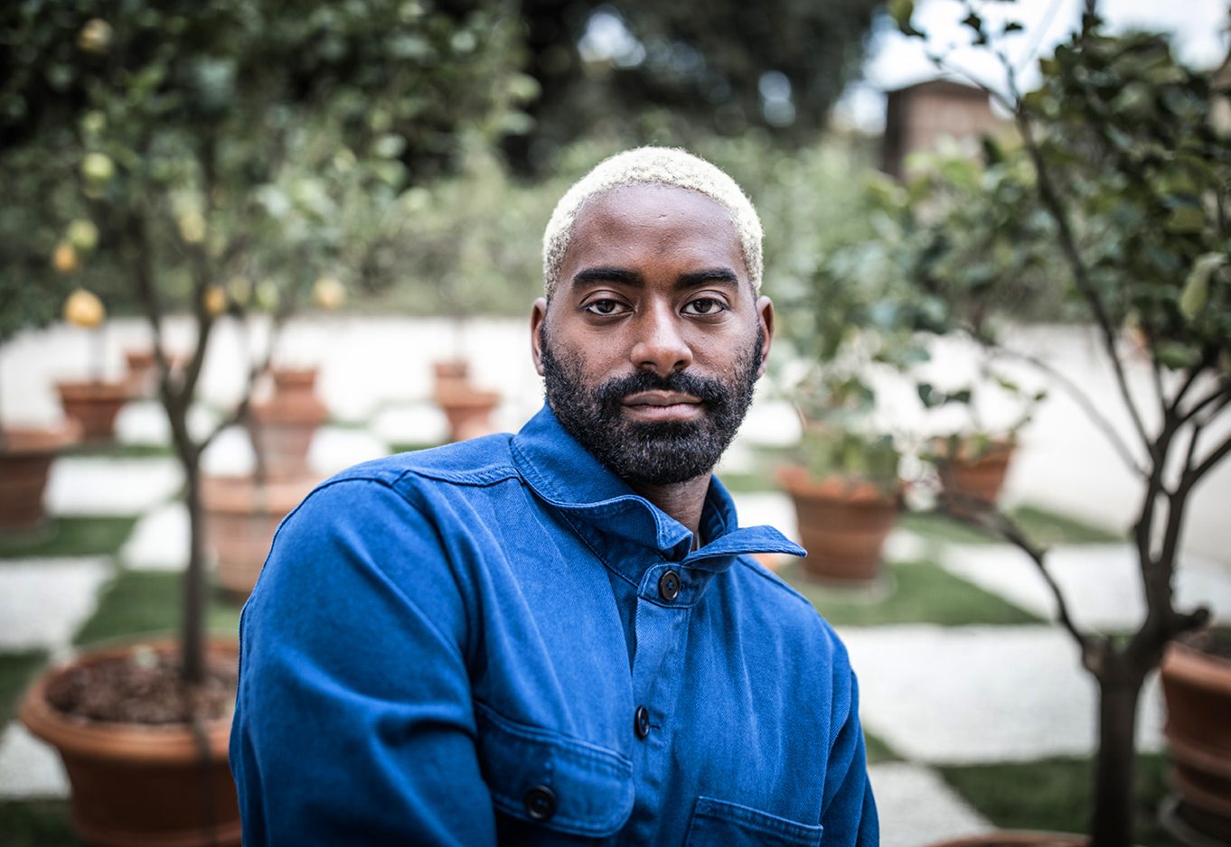 A portrait of Jerron Herman. He is an African American man with a salt and pepper beard and short, bleached hair. He is wearing a blue denim shirt with a pointed collar, sitting at an angle with foliage behind him.