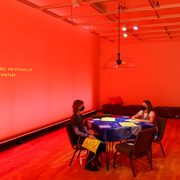 In a gallery, two students sit across from each other at a dining table, with four neon yellow heating pads.The space is entirely filled with a warm red light. On the wall, matching yellow text reads “Our needs and our ways of being together”.