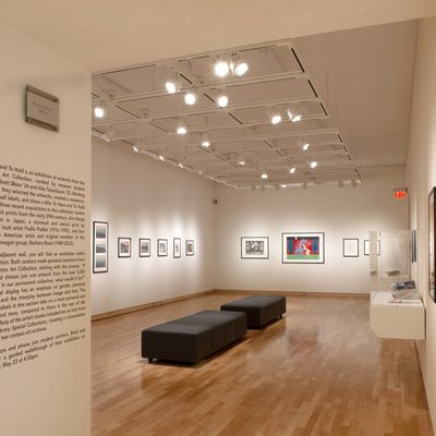 The viewer looks through a doorway into a brightly lit gallery with white walls. Framed art hangs on the walls, and there are two backless black cushioned benches in the center of the room.