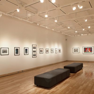 A brightly-lit gallery with white walls. Framed art hangs on the walls, and there are two backless black cushioned benches in the center of the room.