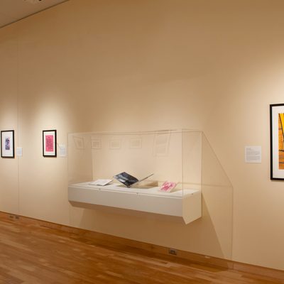 A wall of an art gallery. There are two glass cases containing open books. Three framed pictures hang on the wall as well, one of which depicts a naked chest with hair and breasts.