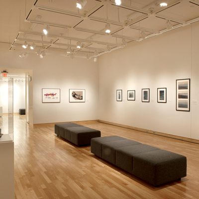 A brightly-lit gallery with white walls. Framed art hangs on the walls, and there are two backless black cushioned benches in the center of the room. Near the viewer, there are two glass cases containing open books.