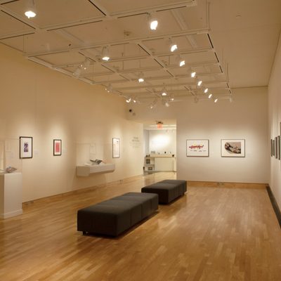 A brightly-lit gallery with white walls. Framed art hangs on the walls, with some in glass cases, and there are two backless black cushioned benches in the center of the room.