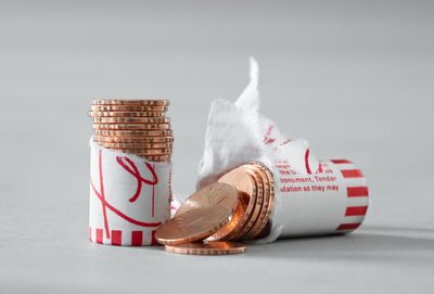 A cracked open roll of pennies which are edge-engraved with the phrase, “THE BODY WAS ALREADY SO FRAGILE”. The image is carefully composed: half of the roll is stacked upright and half is spilling out across a neutral-grey background, which is studio lit.
