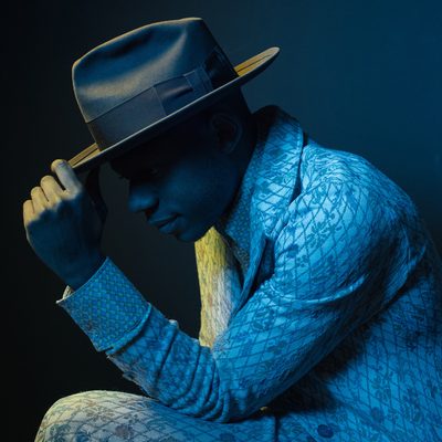 A portrait photograph of musician Ondara in profile facing to the left. His left elbow rests on his left knee, with hand touching the tip of a wide brimmed hat. He wears a light colored suit with a diamond and floral pattern. The image overall is dark blue, skin and suit all taking the blue tone of light, with yellow/green light outlining and highlighting the front of his face and the hand touching the hat. The background is solid darker blue