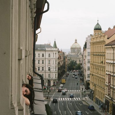 Two girls leaning out of the windows of a building, with a Prague street behind them.