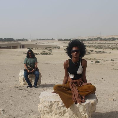 Two people meditating on top of rocks, with a desert and a lake behind them