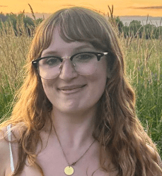 Young white woman with long brown hair and glasses with a field behind her
