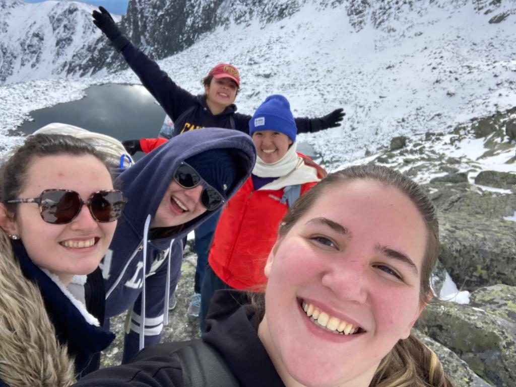 Hiker group selfie with snow-covered rocks and an alpine lake in the background.