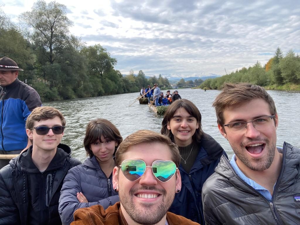 Students in a group selfie on a river with trees lining the banks on either side. Two other rafts and a view of snow-capped mountains can be seen behind them.