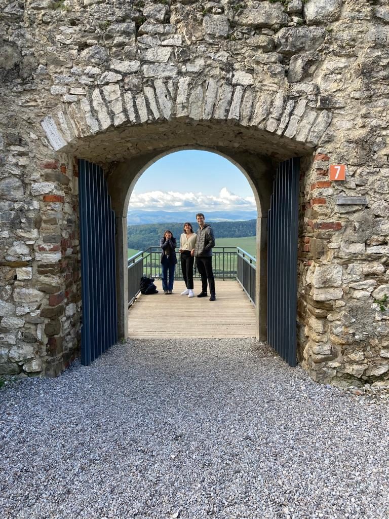 Three students stand far from the camera on a wooden platform with metal railings on either side. A small gated archway in the thick stone wall stands open in front of them. 