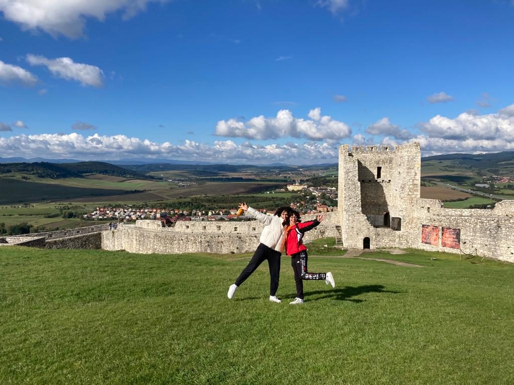 Two students making celebratory poses in the castle courtyard. A tower with a missing wall and a view of the countryside are visible behind them.